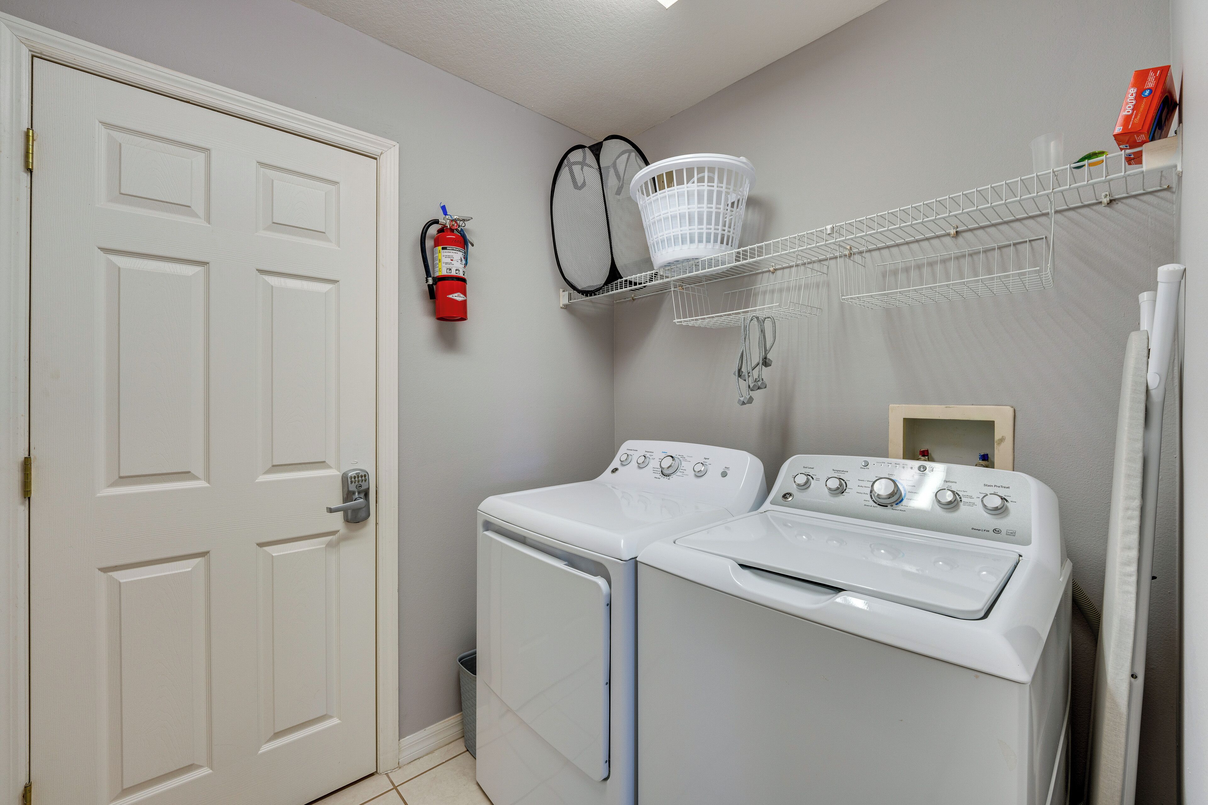 A laundry room with two white front-loading washing machines, a white door, a fire extinguisher, and a shelf with a basket and a box.