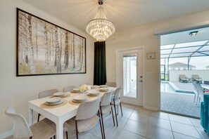 Un coin repas avec une table blanche dressée pour six personnes, un lustre, un grand tableau sur le thème de la forêt et une vue sur une piscine extérieure, visible à travers une porte vitrée.