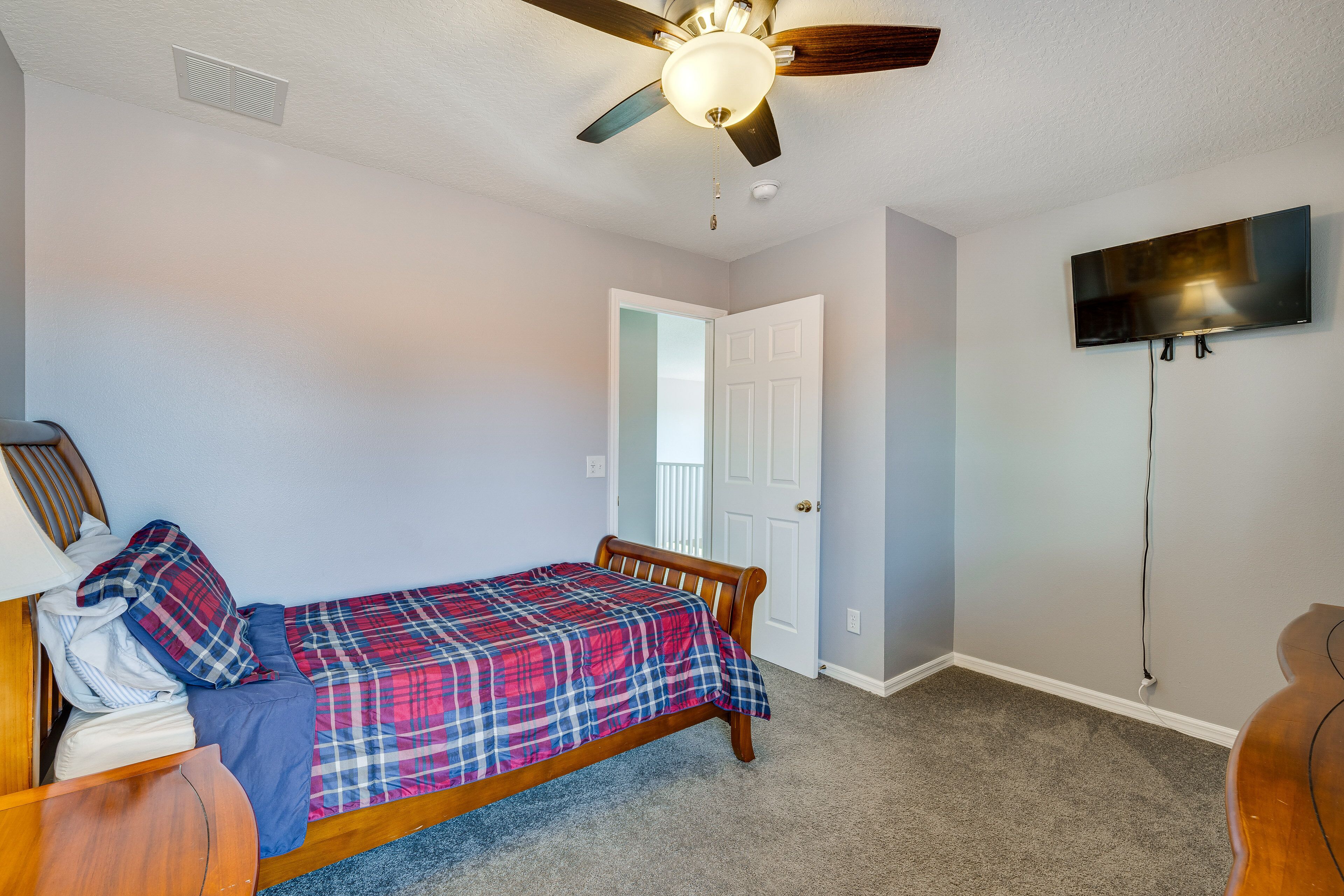 A bedroom with a bed, a ceiling fan, a TV mounted on the wall, and a door leading to another room.
