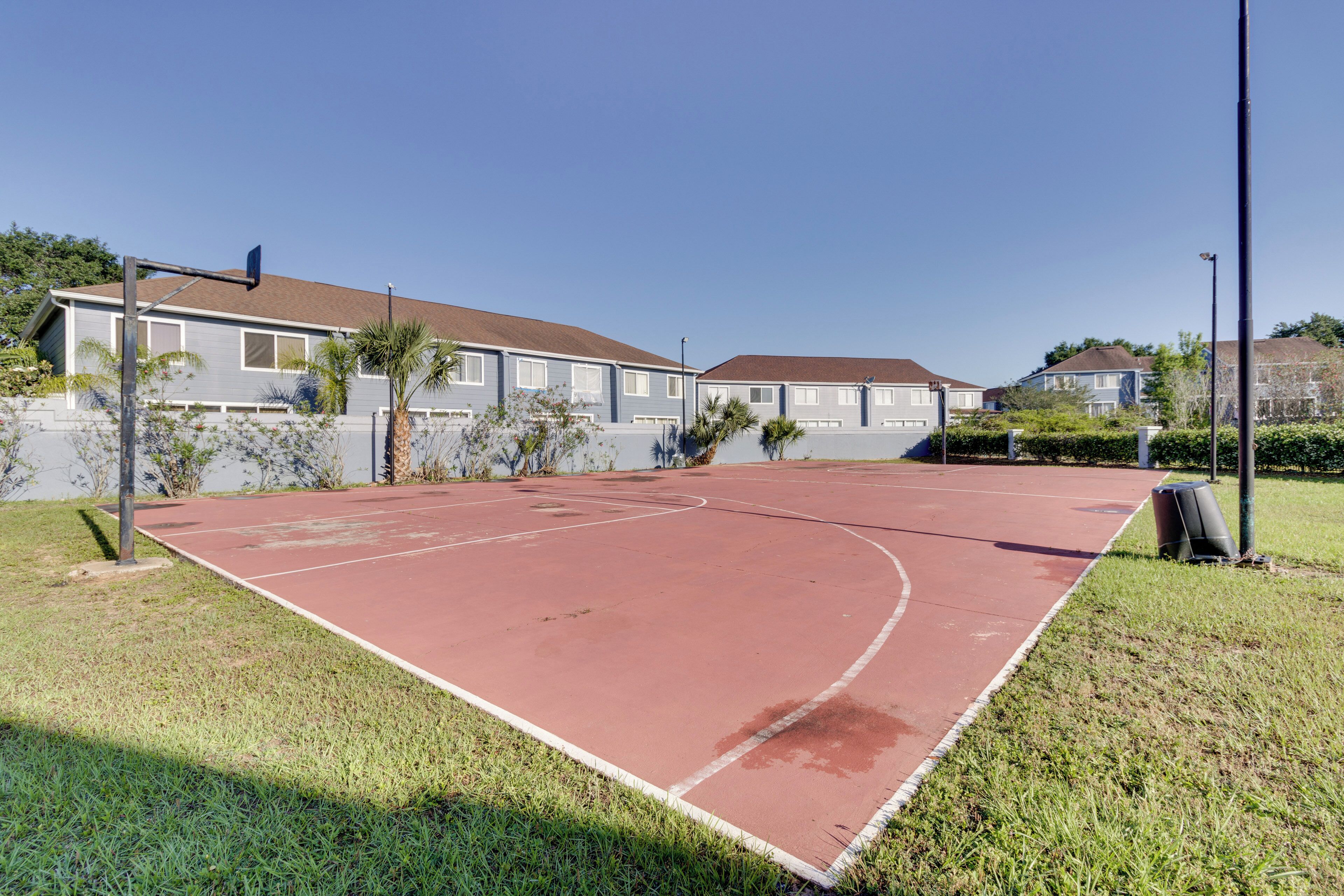 Un terrain de basket en plein air, doté d’une surface rouge, entouré de pelouse et de palmiers, avec des maisons en arrière-plan.