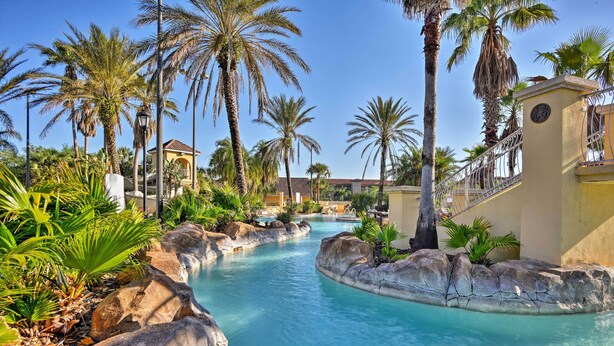 A pool area with palm trees, rocks, and a building with a clock. - 9 Mi to Disney: Regal Palms Townhome w/ Patio! (Davenport)