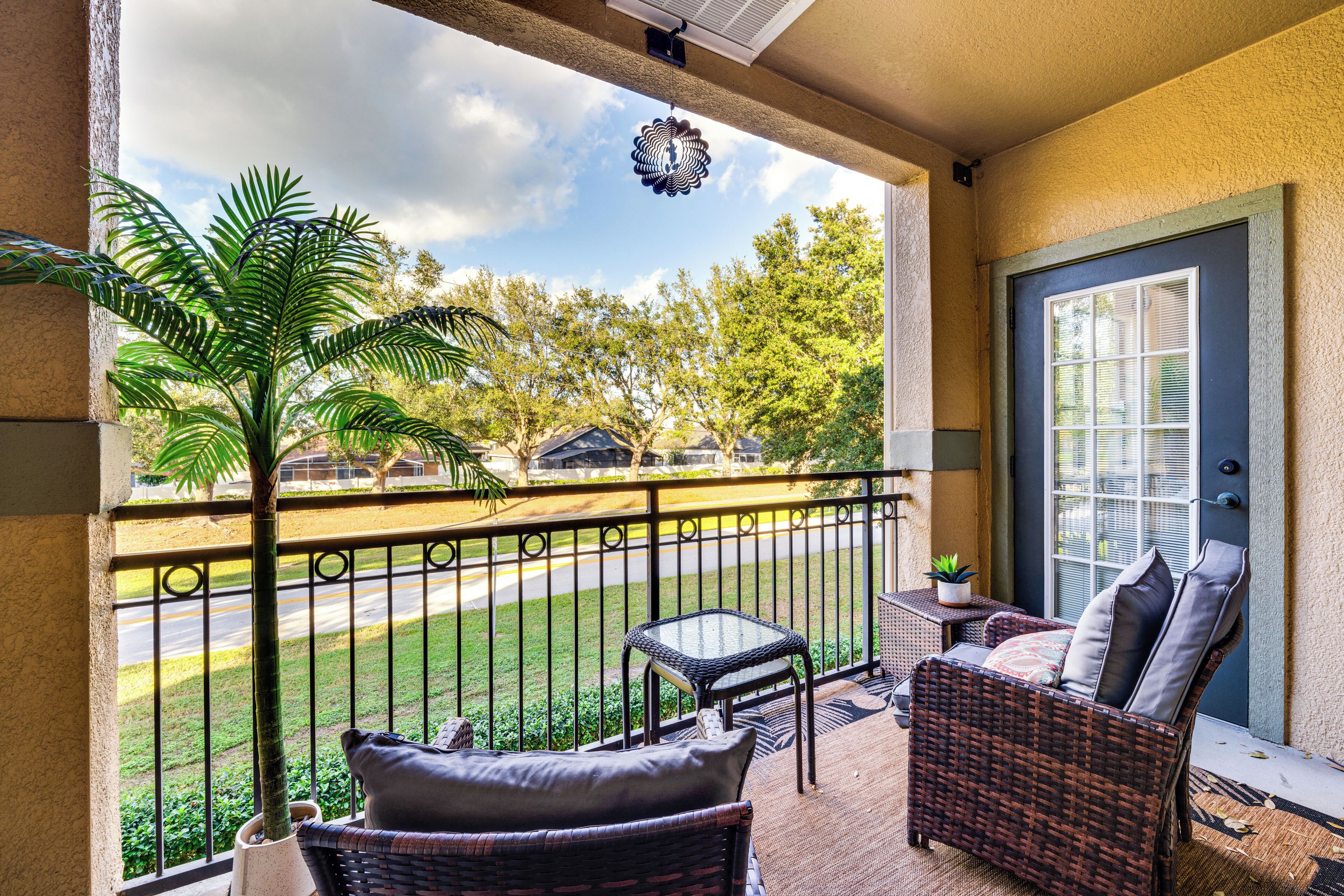 A balcony with wicker furniture, a potted plant, and a view of a street and trees.