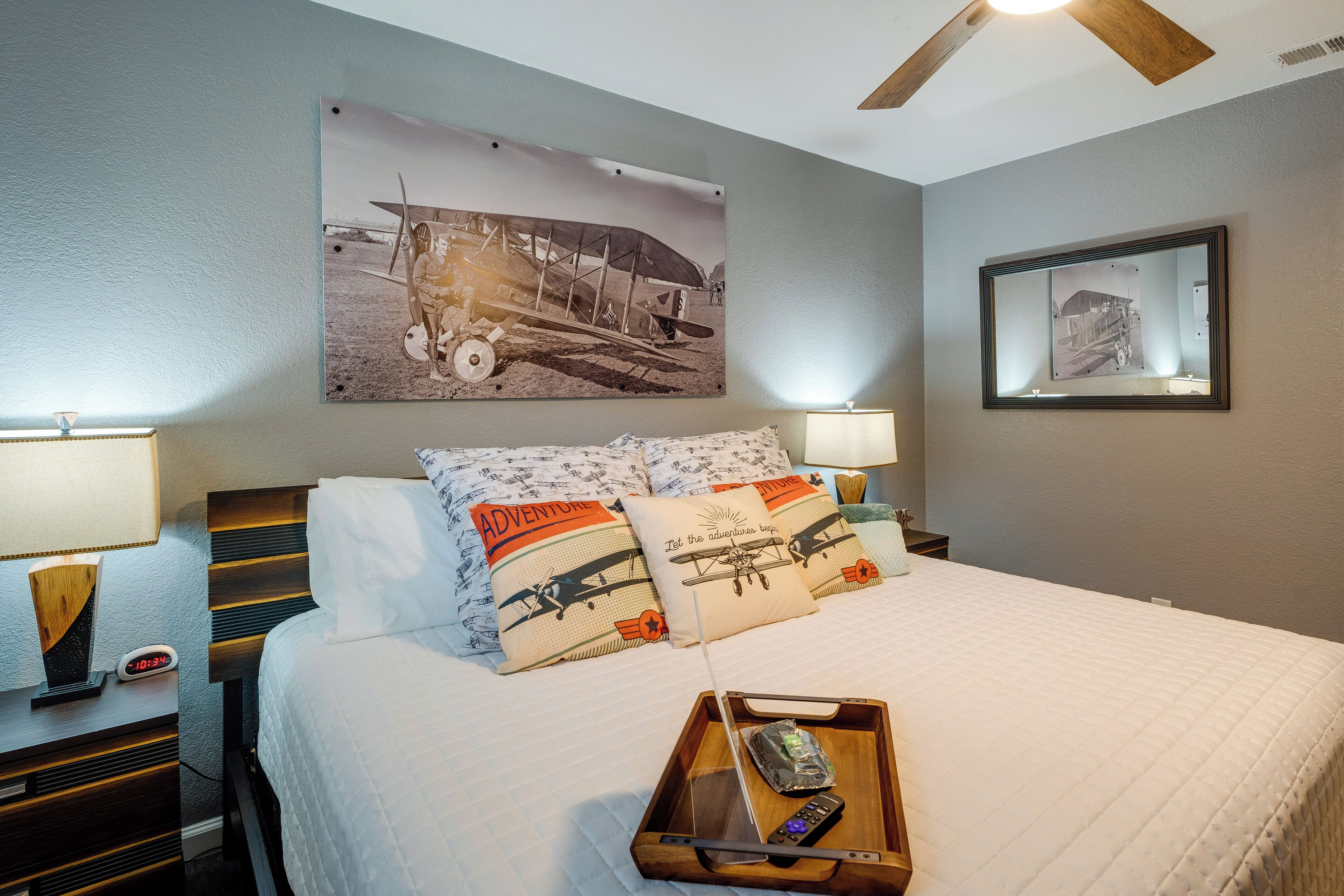 A neatly made bed with patterned pillows, a tray with a remote control, and a wall-mounted airplane picture.