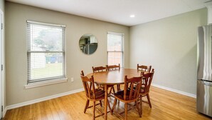Un coin repas avec une table ronde en bois et des chaises, deux fenêtres équipées de stores, un miroir rond accroché au mur et un réfrigérateur en acier inoxydable.