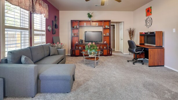 A living room with a grey sectional sofa, a glass coffee table with decorative items, a wooden entertainment unit with a flat-screen TV, a wooden desk with a chair, and a ceiling fan.