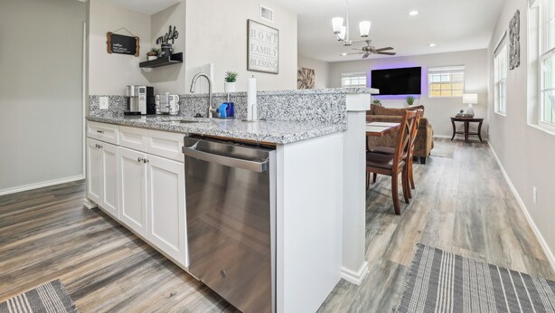 A modern kitchen with white cabinets, a stainless steel dishwasher, granite countertops, and a dining area with wooden chairs and a TV. - Spacious San Antonio Retreat w/ Private Patio! (San Antonio)