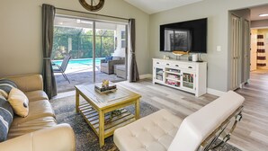 A living room with a sofa, coffee table, TV, and a sliding glass door leading to a pool area.