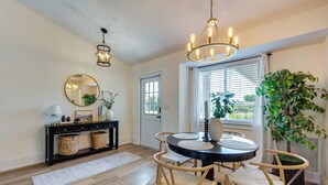 Un coin repas comprenant une table ronde, quatre chaises, un lustre, une console avec des paniers, un miroir et une plante en pot.