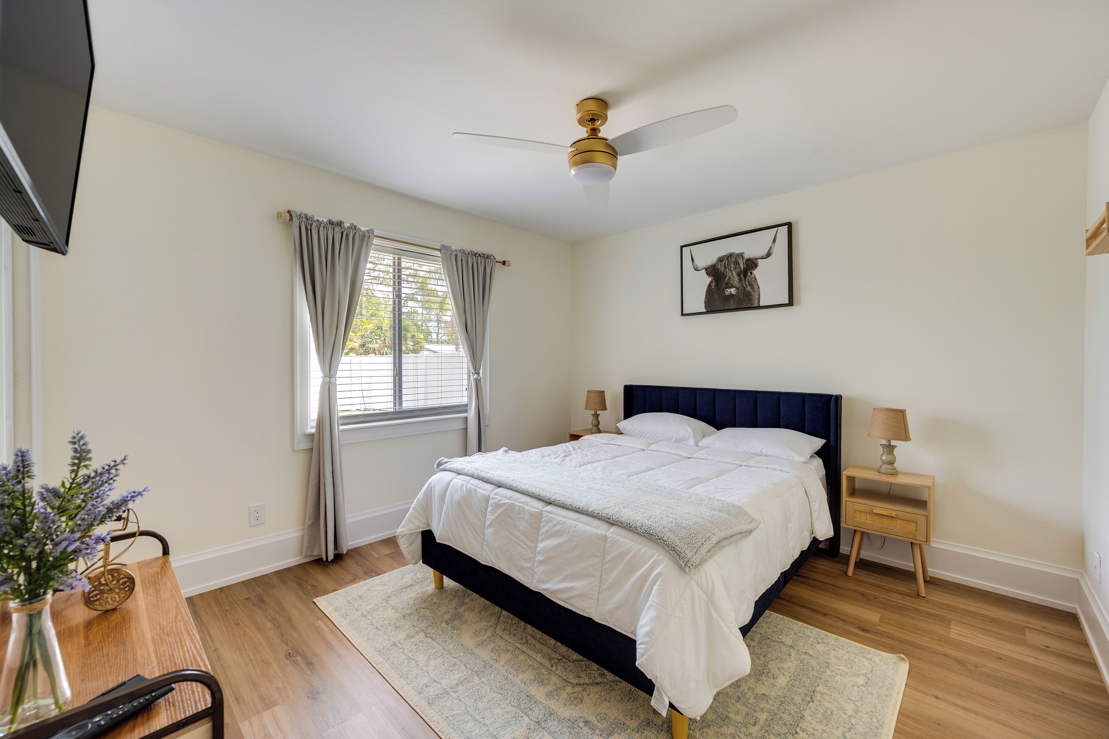 Une chambre à coucher comprenant un lit, des lampes de chevet, un ventilateur de plafond, une fenêtre avec des rideaux, un téléviseur fixé au mur et un tableau encadré.