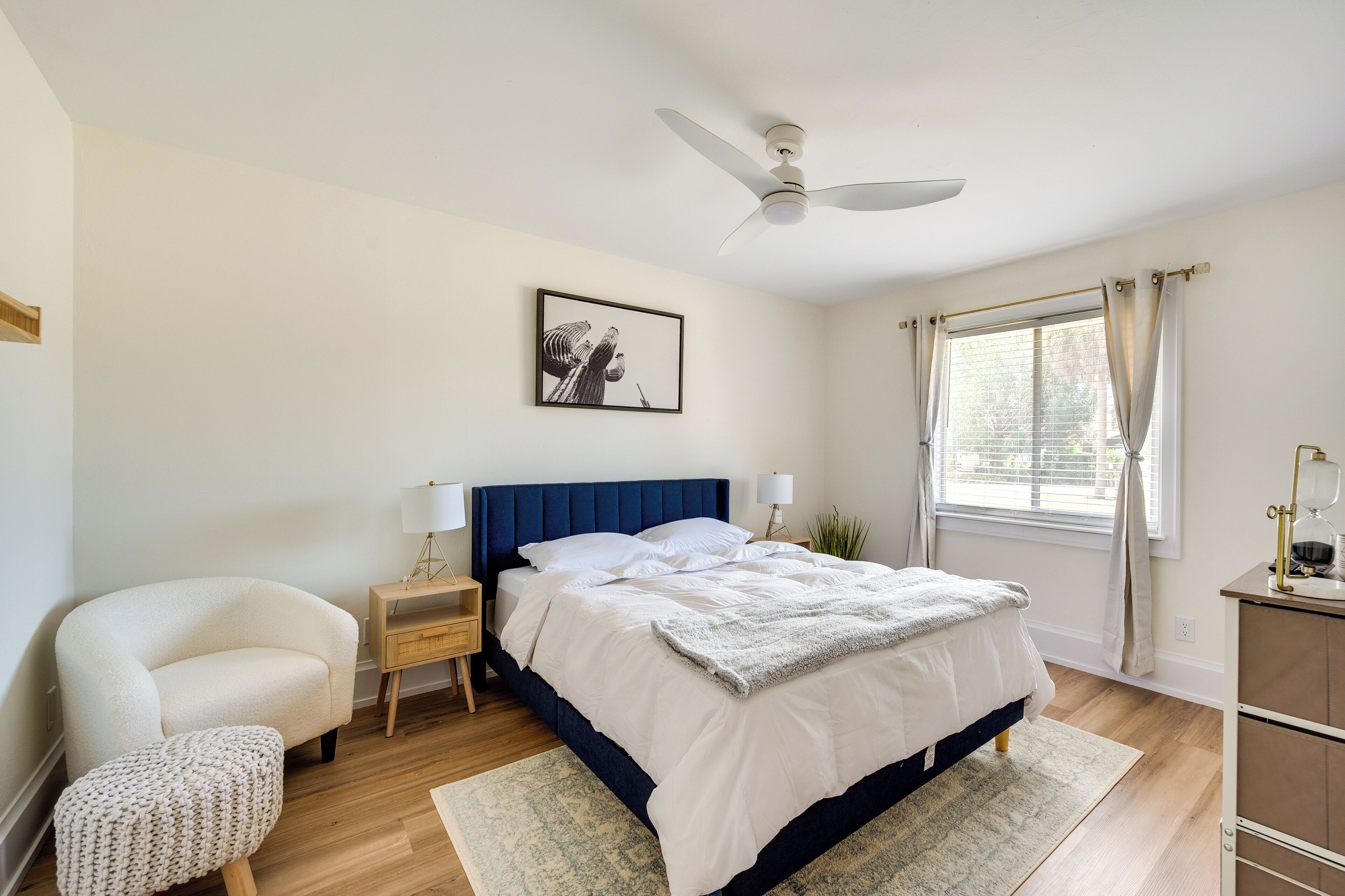 Une chambre à coucher comprenant un lit, une chaise, une table de chevet, une lampe, un ventilateur de plafond, une fenêtre avec des rideaux et une plante.