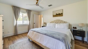 Une chambre à coucher comprenant un lit, une table de chevet, une lampe, un ventilateur de plafond et une fenêtre avec des rideaux.