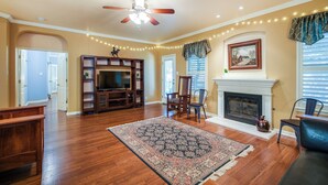 A spacious living room with hardwood floors, a ceiling fan, a fireplace, and a large entertainment center. - North San Antonio Vacation Rental w/ Yard & Deck! (San Antonio)