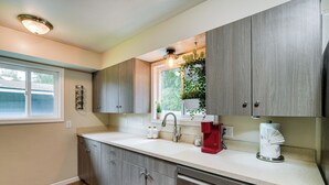 A modern kitchen with wooden cabinets, a white countertop, a sink, a coffee maker, and a window with plants. - Anchorage Home ~ 7 Mi to Chugach State Park! (Anchorage)