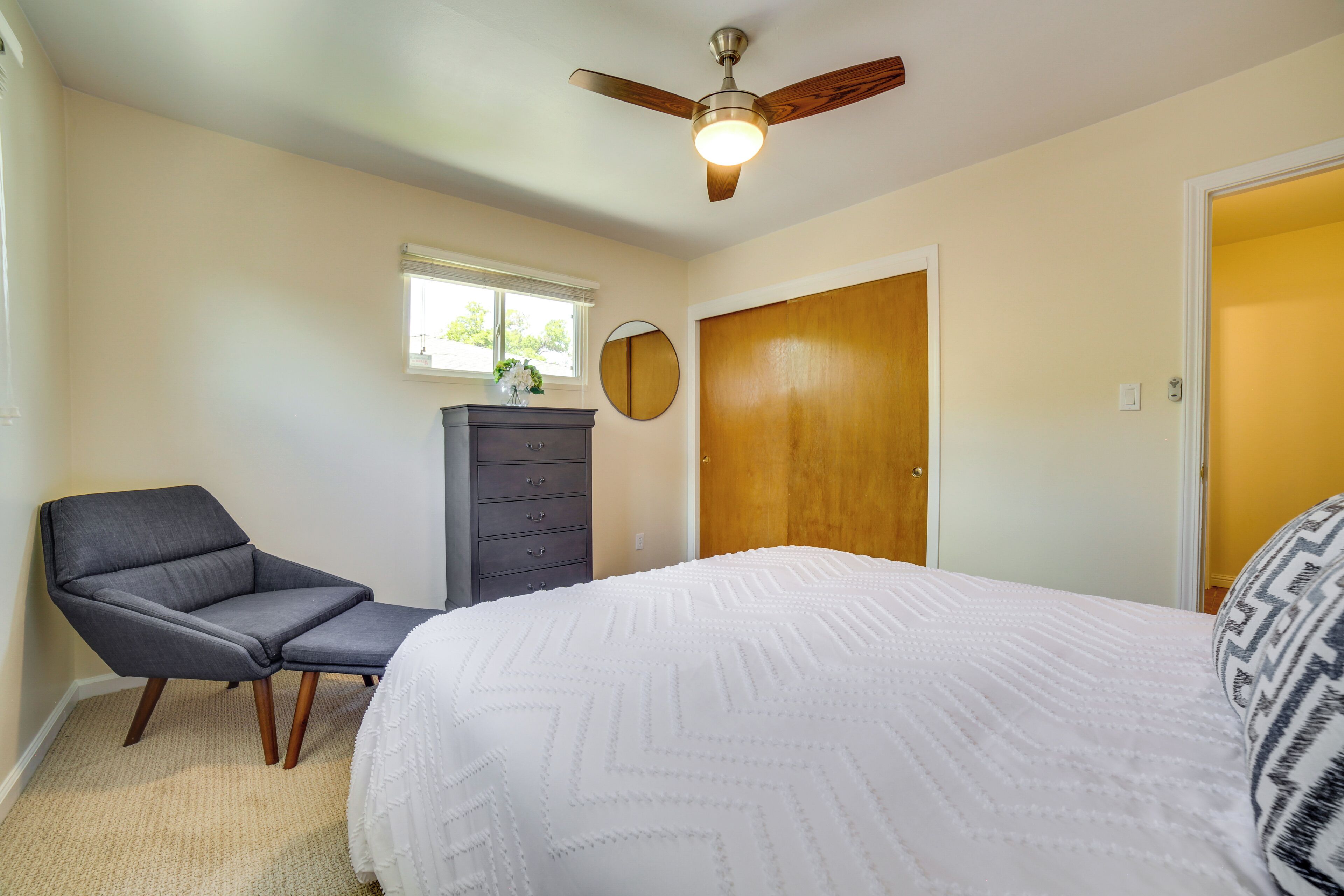 A bedroom with a bed, a chair, a dresser, a ceiling fan, a window, and a door.