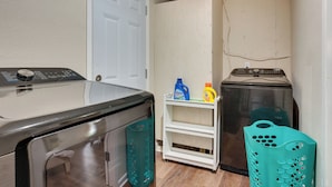 A laundry area with a washer and dryer, a white shelf with cleaning supplies, and a teal laundry basket.