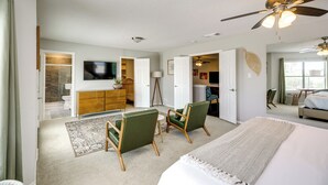 Une chambre spacieuse avec un grand lit, deux fauteuils verts, un ventilateur de plafond et un téléviseur fixé au mur.