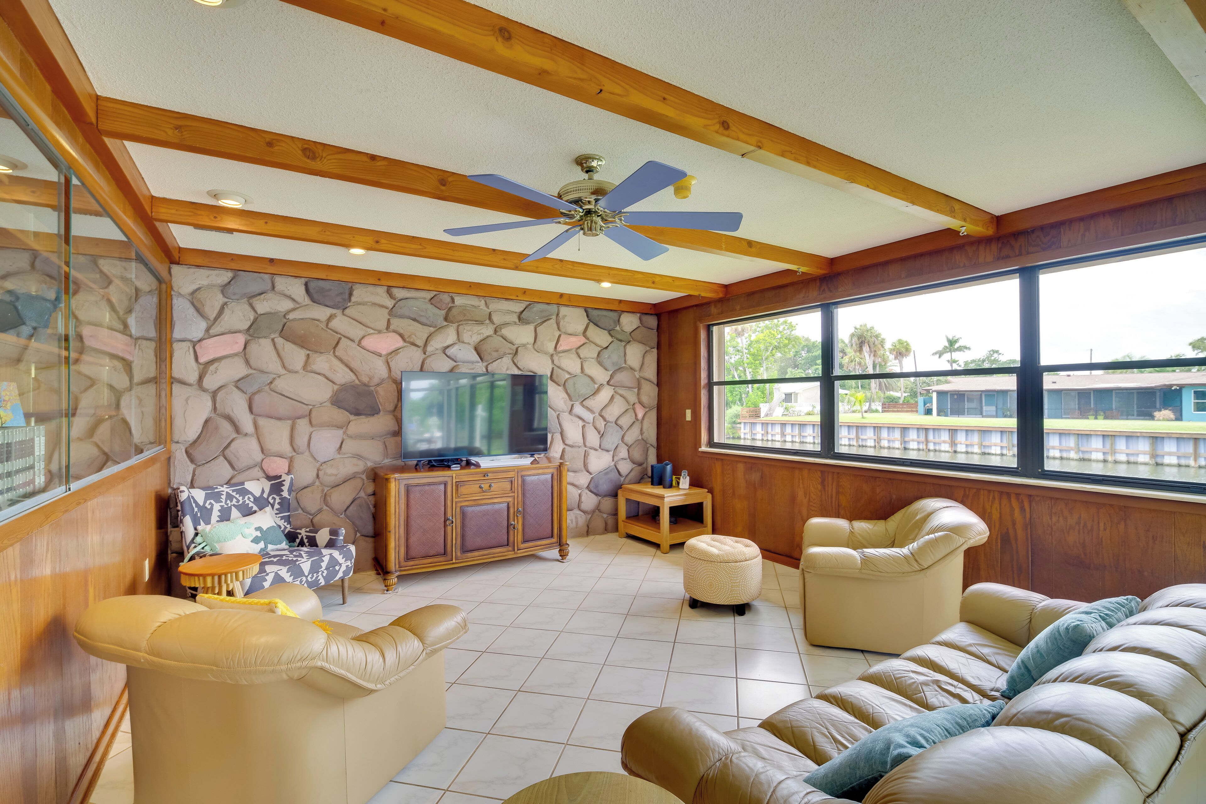 Un salón espacioso con pared de piedra, vigas de madera en el techo, ventilador de techo, televisor y ventanas grandes que ofrecen vistas al exterior.