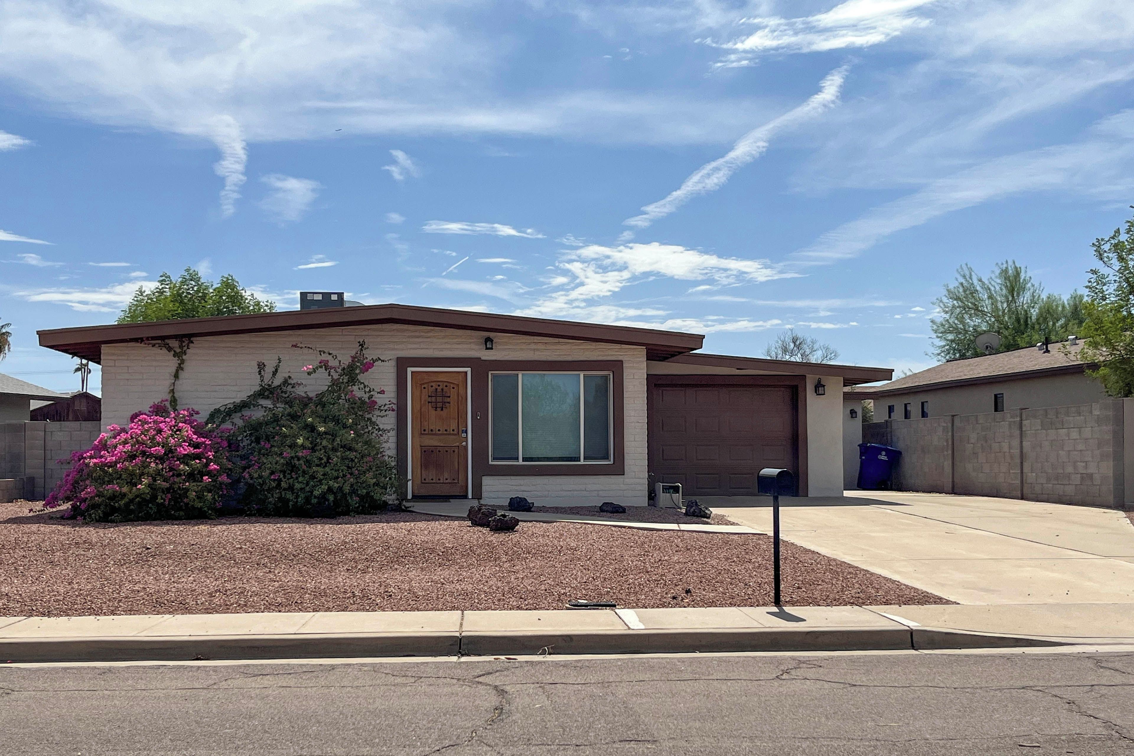 A single-story house with a garage, a front yard with mulch and a flowering shrub, and a clear sky.