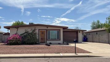 A single-story house with a garage, a front yard with mulch and a flowering shrub, and a clear sky.