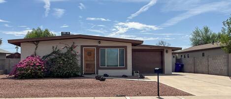 A single-story house with a garage, a front yard with mulch and a flowering shrub, and a clear sky.