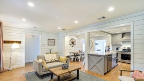 A spacious living room with a sofa, coffee table, and a dining area with chairs. A kitchen with modern appliances is visible in the background. - Pecan Blue Frisco Hub: Dtwn, Dining & Stadiums! (Frisco)