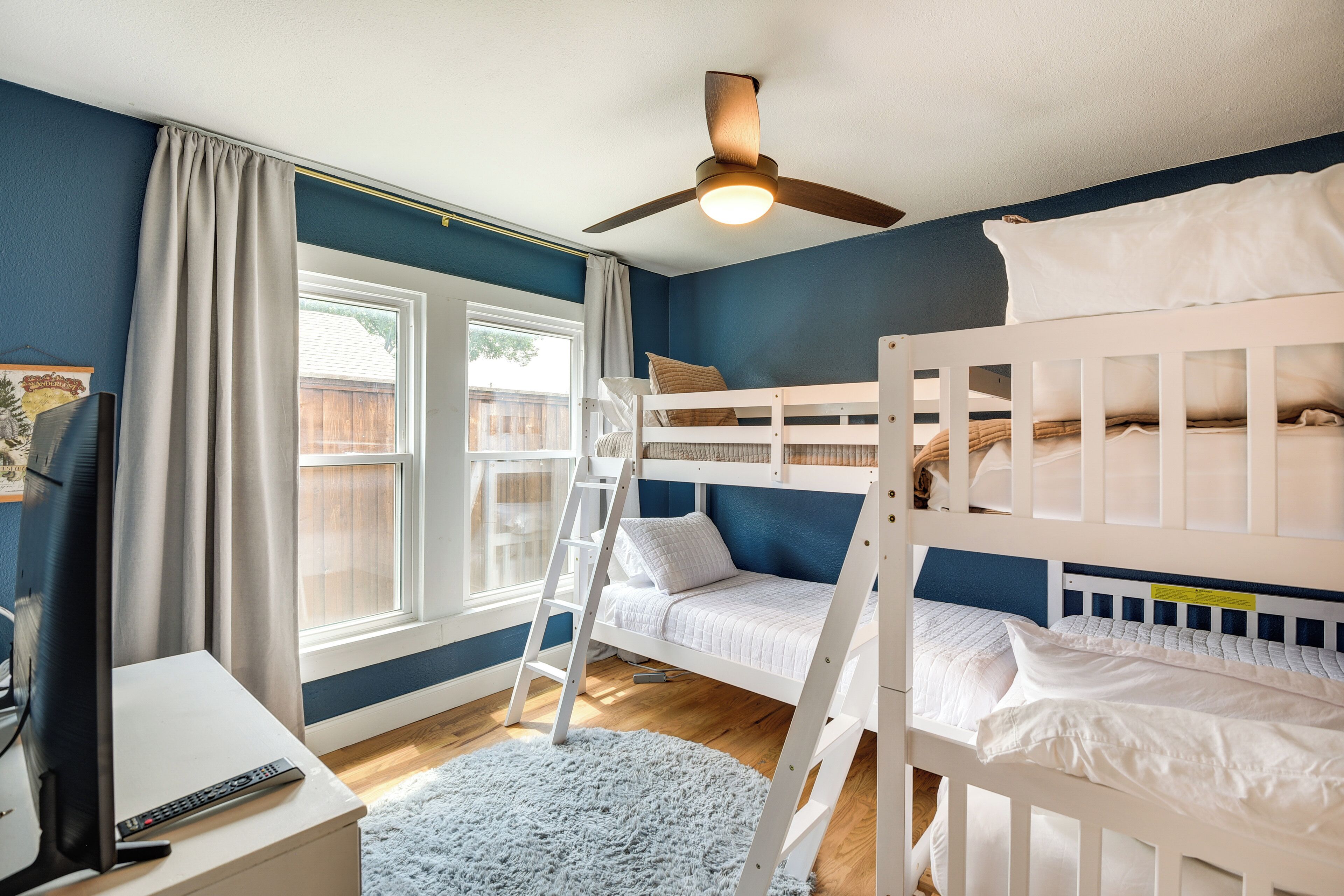 A room with bunk beds, a ceiling fan, a window with curtains, a wooden floor, and a remote control on a table.