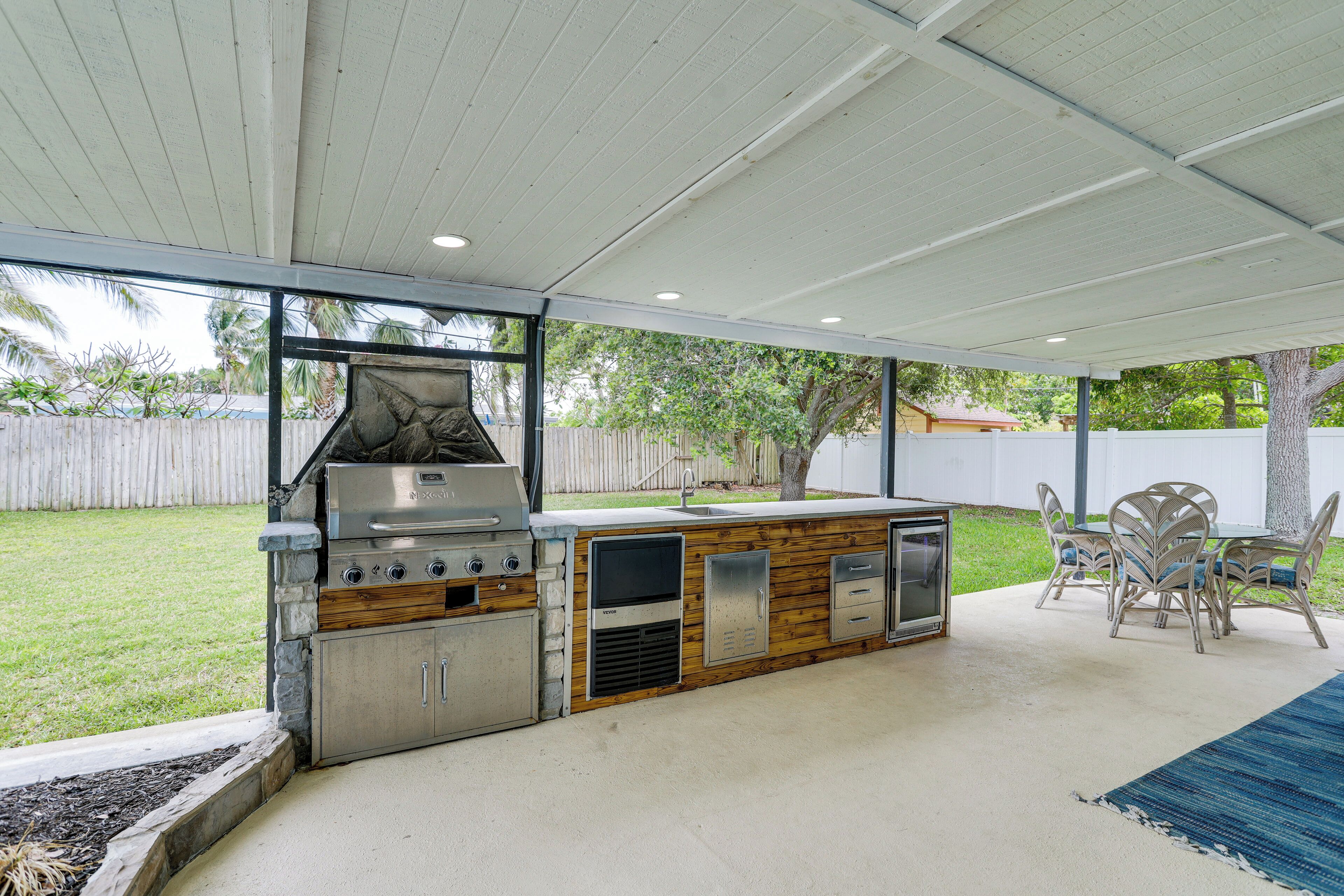 An outdoor kitchen with a grill, sink, and storage cabinets, surrounded by a covered patio area with a table and chairs.