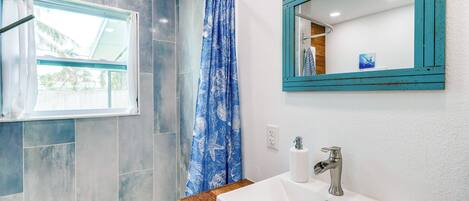 A bathroom with a shower, a window, a sink, and a mirror.