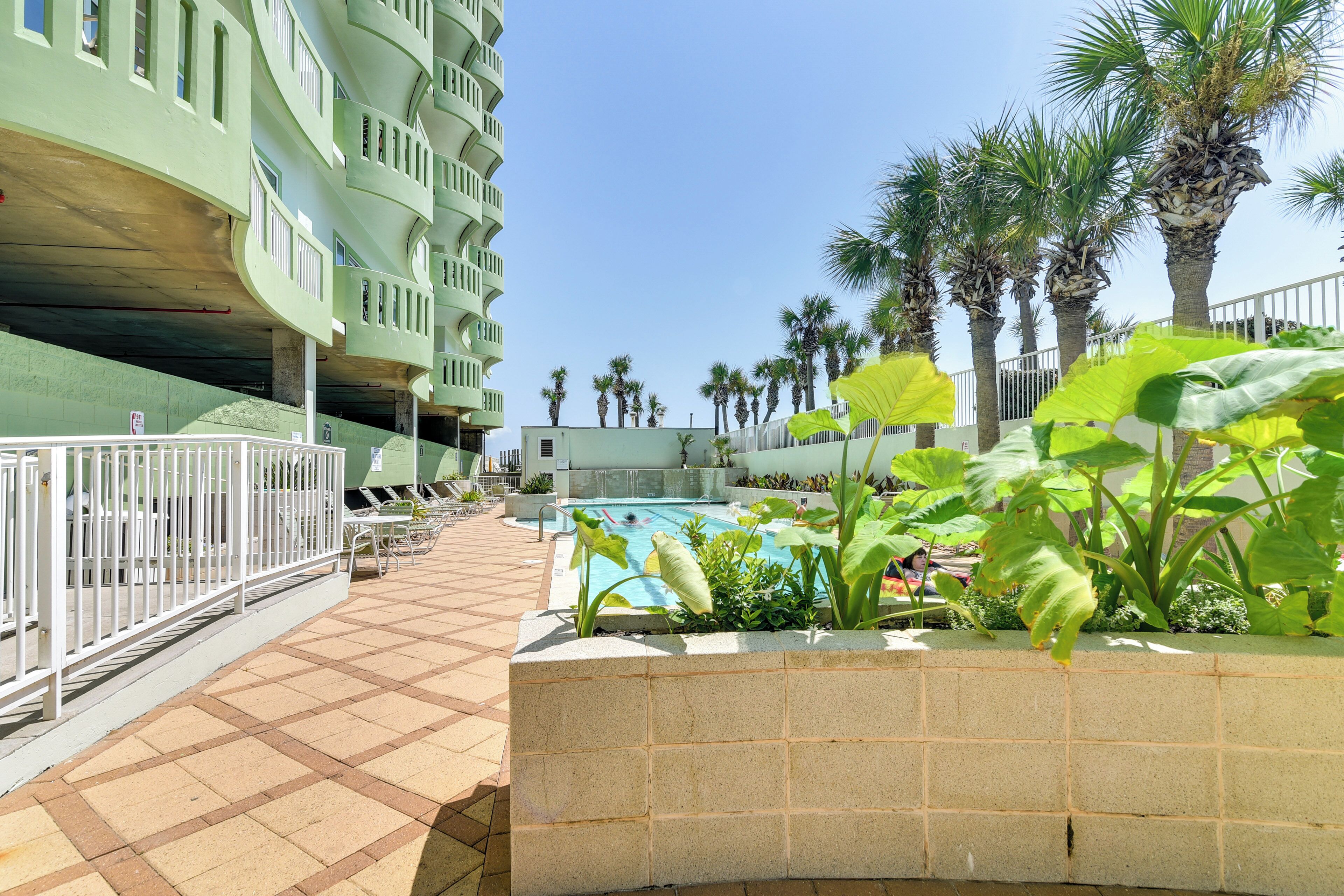 A pool area with a tiled floor, surrounded by palm trees and a building with balconies.