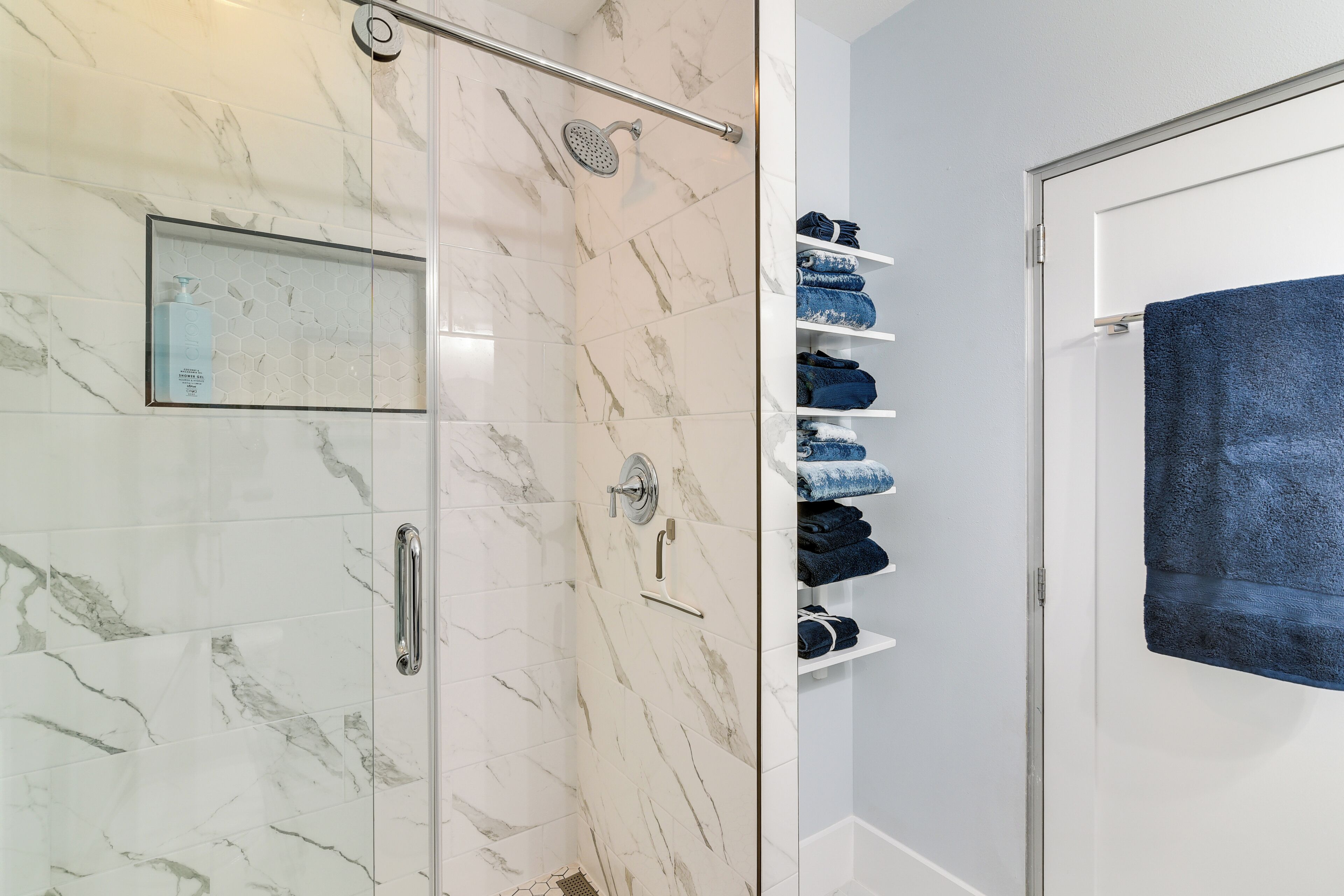 A bathroom with a glass shower enclosure, marble tiles, a towel rack with folded towels, and a blue towel hanging on a rack.