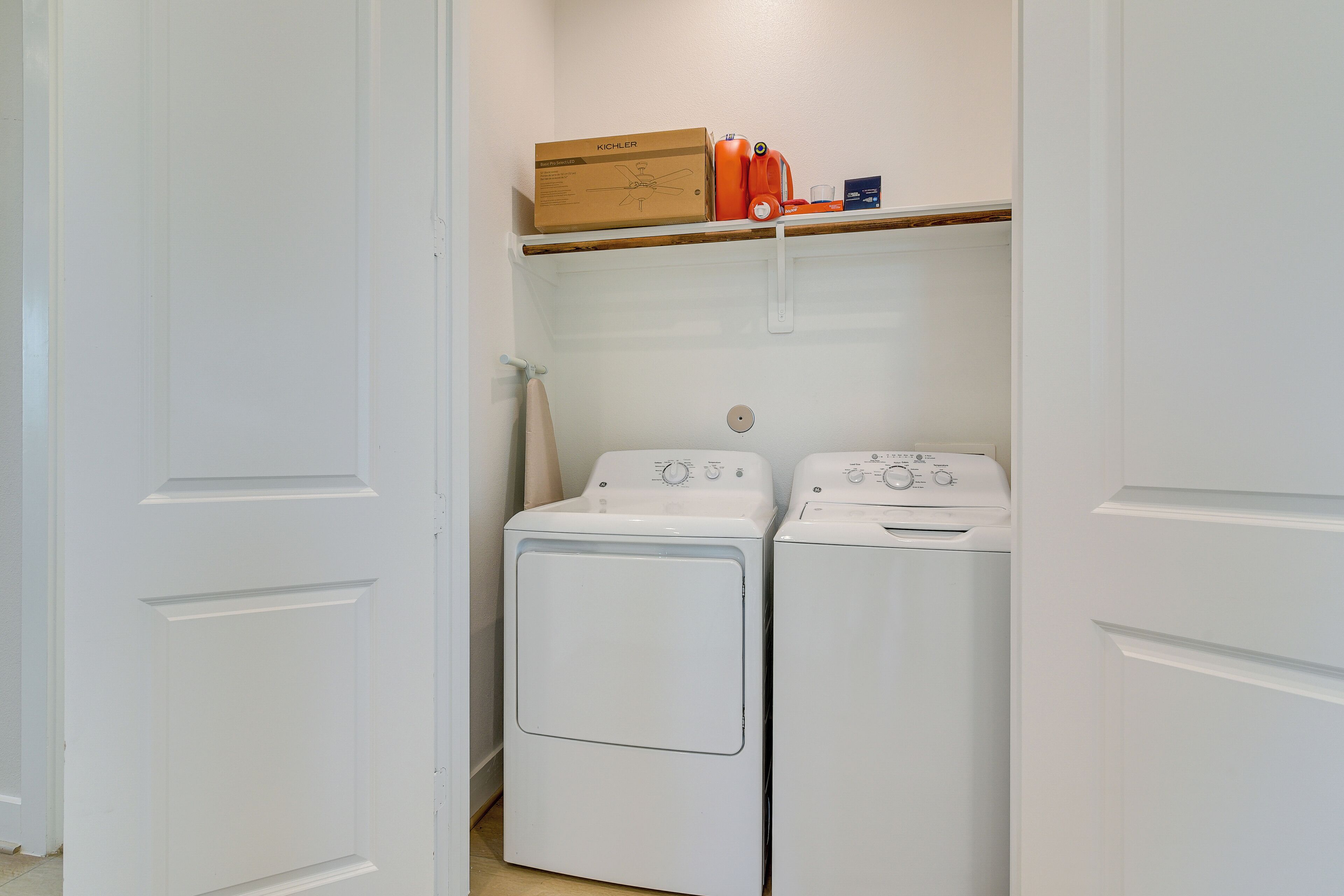 A laundry room with two white front-loading washing machines and a shelf above containing a box, a bottle, and a container.