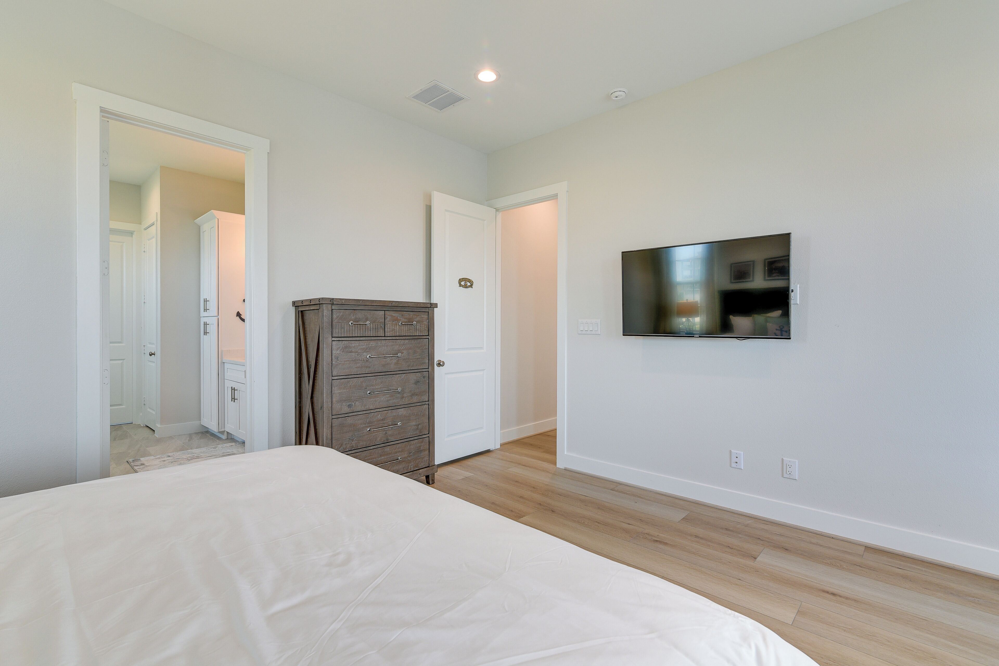 Un dormitorio con cama, cómoda, televisor montado en la pared y puerta que da a otra habitación.