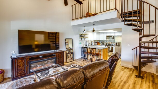 A spacious living room with a large flat-screen TV, leather sofa, and a wooden entertainment unit. A staircase leads to an upper level with a balcony. - Chandler Vacation Rental w/ Pool & Hot Tub Access (Chandler)
