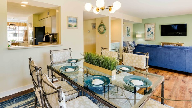 Un salon moderne avec un canapé bleu, un coin repas avec une table et des chaises en verre, et une cuisine avec des meubles blancs et un réfrigérateur noir.