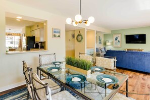 Ein modernes Wohnzimmer mit einem blauen Sofa, einem Essbereich mit einem Glastisch und Stühlen sowie einer Küche mit weißen Schränken und einem schwarzen Kühlschrank.