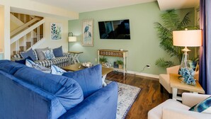 Un salon avec un canapé bleu, une table basse en bois et un fauteuil blanc. On aperçoit un escalier au fond, un tableau au mur et une plante en pot près de la fenêtre.