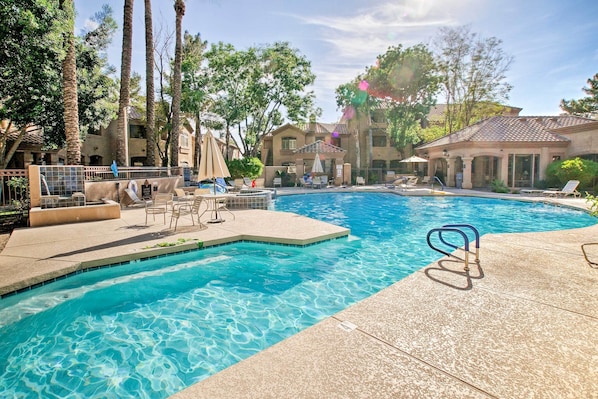 A swimming pool with lounge chairs and umbrellas, surrounded by palm trees and buildings.