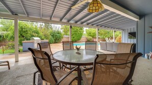 A covered patio with a round table and chairs, a ceiling fan, and a view of a backyard with a grill and pool.