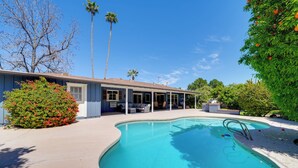 A house with a swimming pool, a covered patio, and a garden with a palm tree and a bush with red flowers.