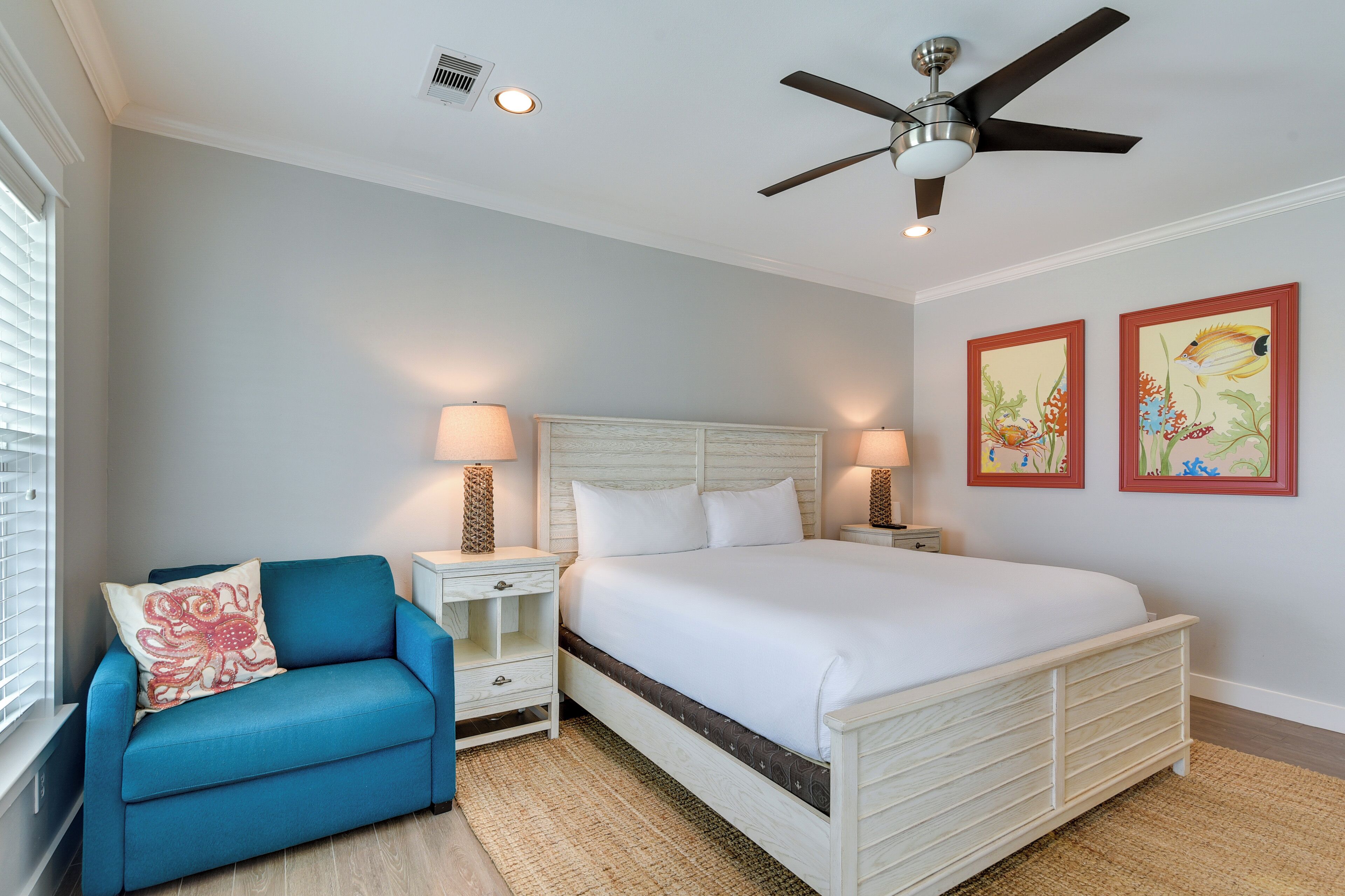 A bedroom with a bed, nightstands, a blue chair, a ceiling fan, and two wall-mounted paintings.