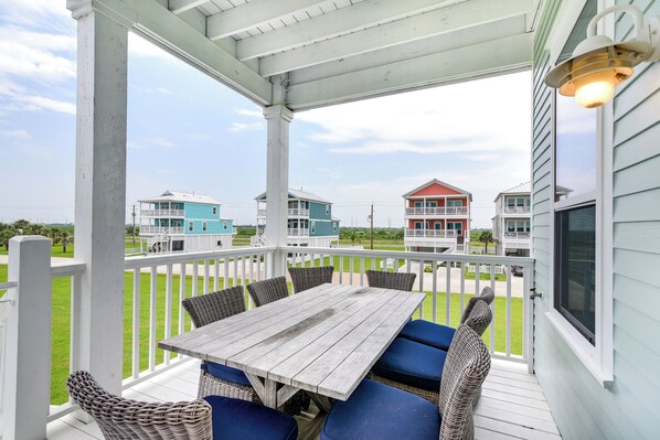 A covered outdoor area with a wooden table and blue cushioned chairs, overlooking a grassy area with colorful houses in the background.