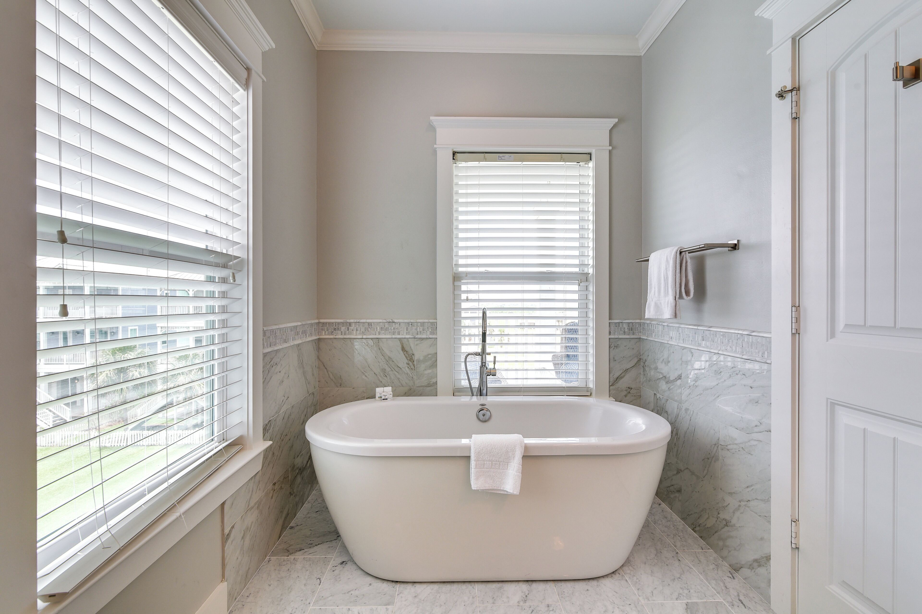 Ein modernes Badezimmer mit freistehender Badewanne, großem Fenster mit Jalousien und Marmorboden.