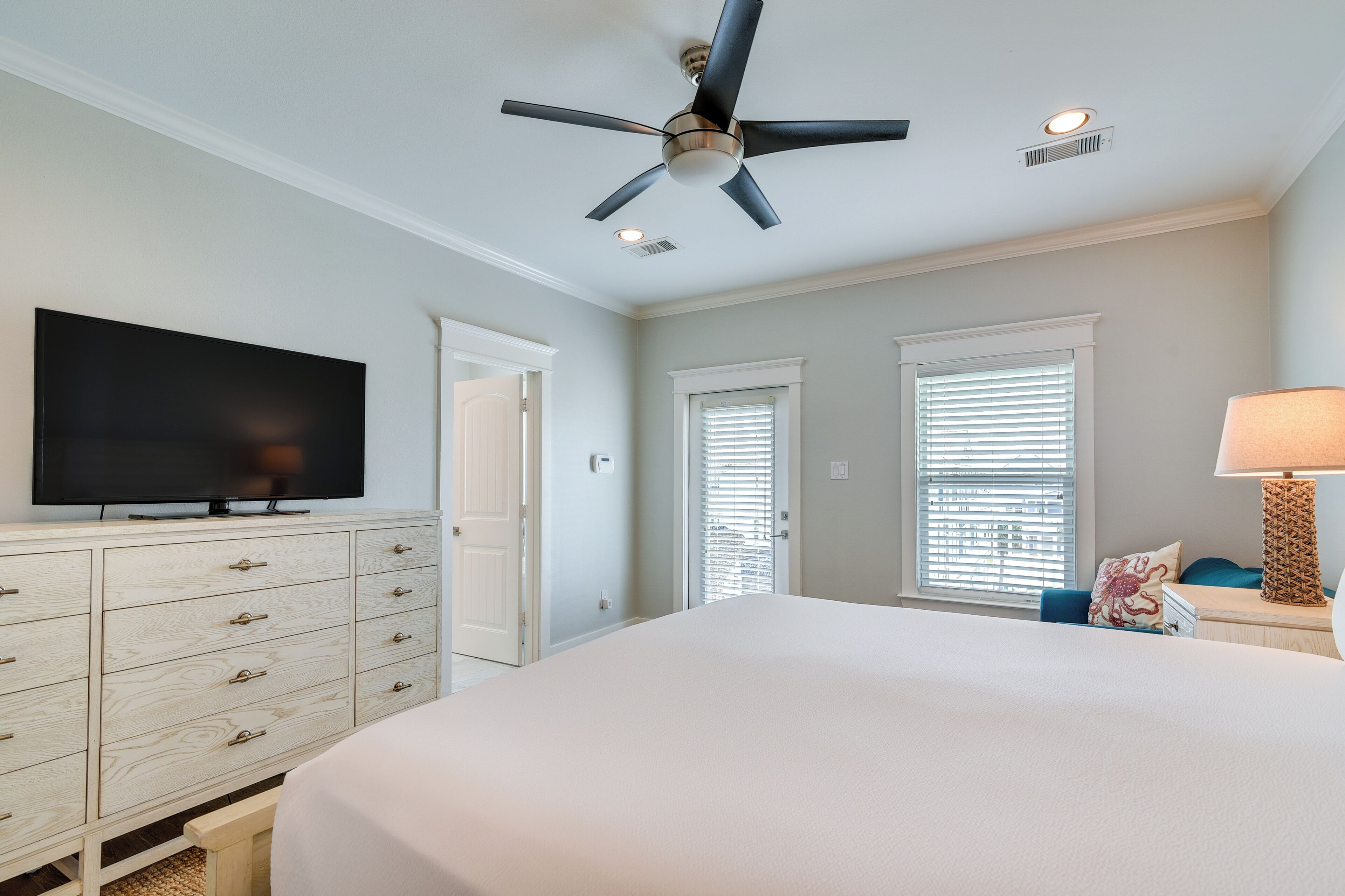 Une chambre à coucher avec un lit, un téléviseur posé sur une commode, un ventilateur de plafond et une fenêtre avec des stores.