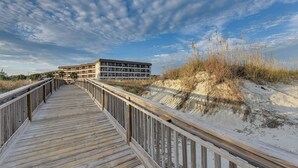 A wooden boardwalk leading to a multi-story building with a flat roof, surrounded by sand dunes and sparse vegetation. - Top-floor Condo w/ Ocean Views in Hilton Head! (Hilton Head Island)