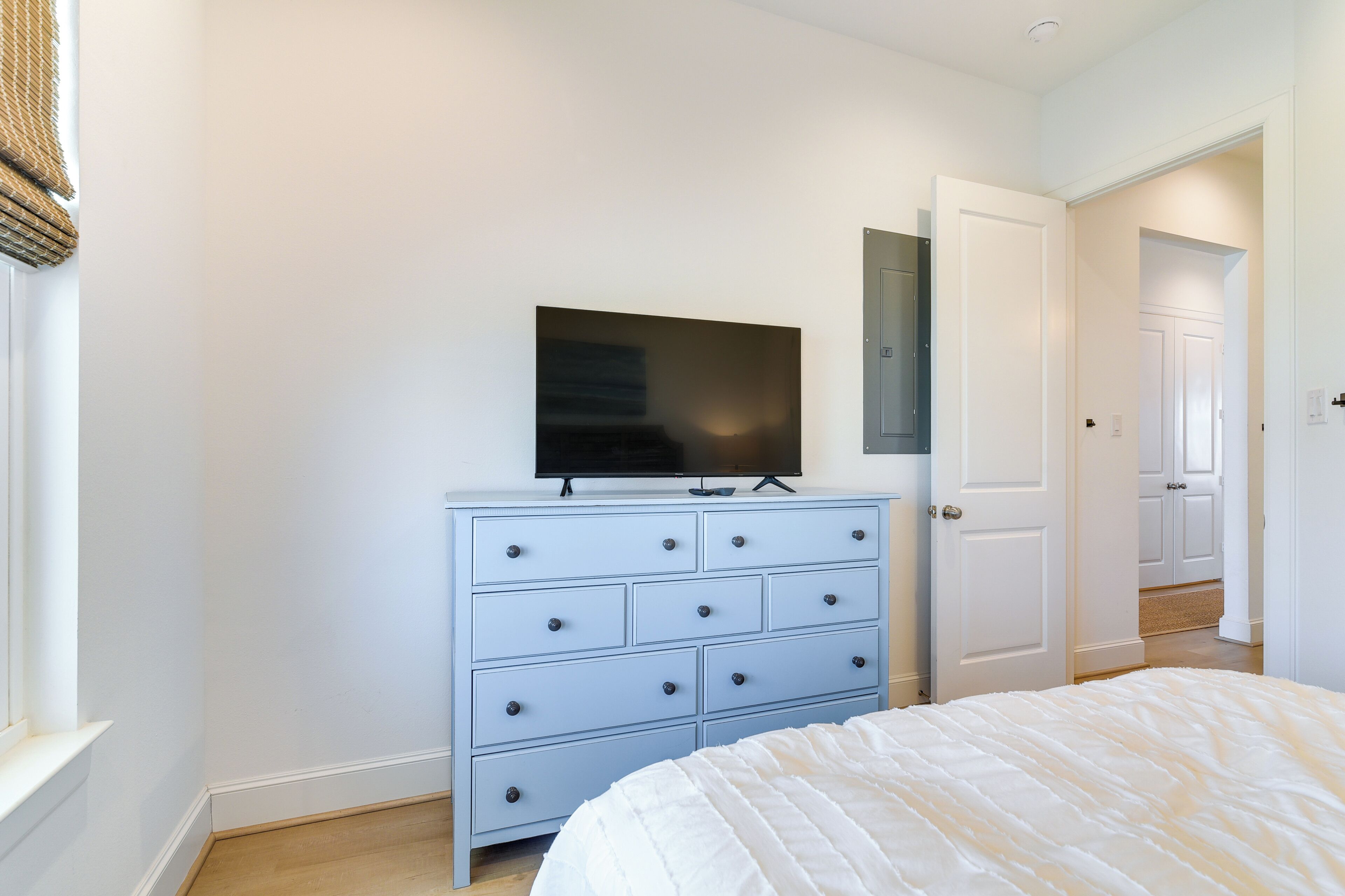Une chambre à coucher avec un lit, une commode sur laquelle est posée une télévision, et une porte qui donne sur une autre pièce.