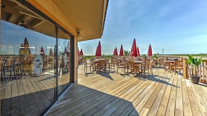 An outdoor wooden deck with tables and chairs, red umbrellas, and a glass wall reflecting the interior. - Hilton Head Island, Beach Resort Condo w/ Pools! (Hilton Head Island)