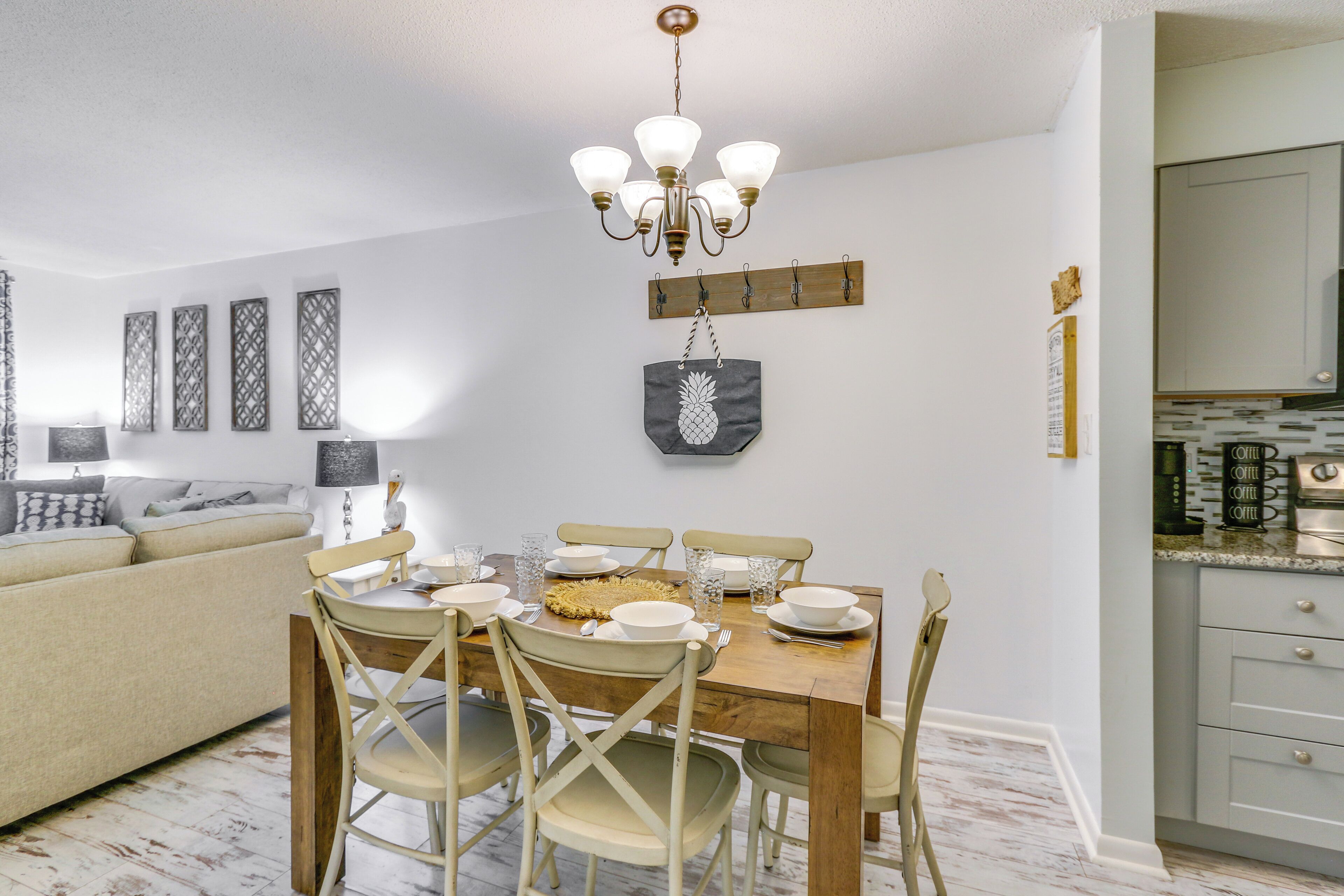 A dining area with a wooden table set for four, surrounded by beige chairs. A chandelier hangs above the table, and a wooden board with hooks is mounted on the wall.