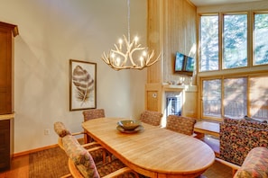 A dining area with a wooden table, floral-patterned chairs, a chandelier, a framed leaf artwork, and a television on the wall. - Pet-friendly Park City Condo w/ Private Hot Tub! (Park City)