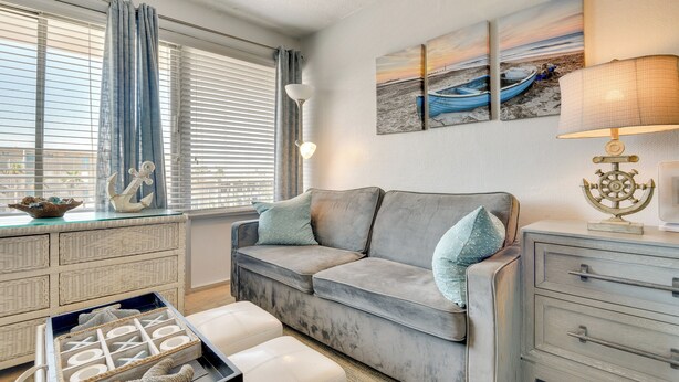 Un salon avec un canapé gris, une petite table sur laquelle se trouve une lampe, et des œuvres d’art représentant une scène de plage.