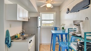 Une cuisine compacte avec des meubles blancs, une cuisinière et un ventilateur de plafond. On y trouve une fenêtre avec des stores, un évier et une table bleue avec des chaises.